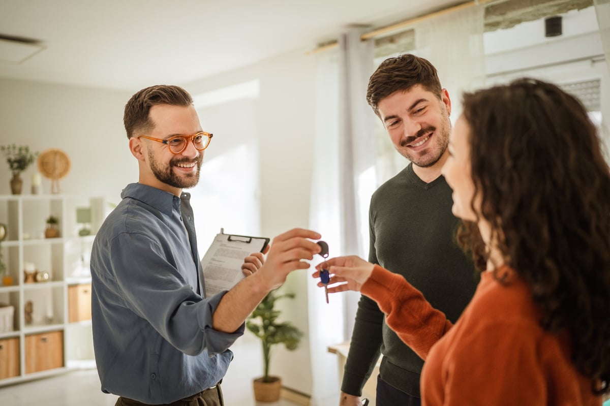 Real estate agent smiling while handing over new house keys to a happy young couple, celebrating their property ownership and successful moving into their modern apartment